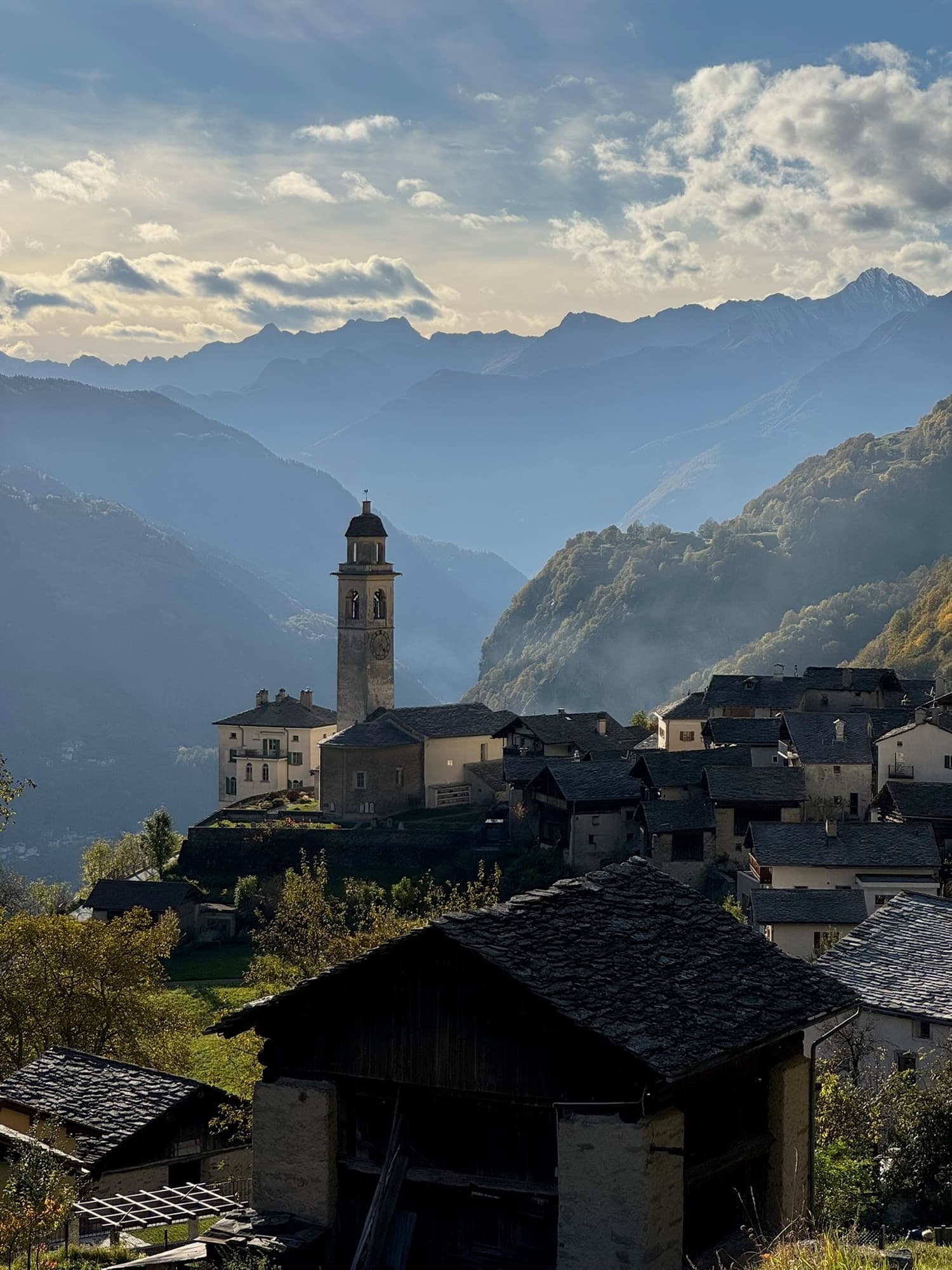 Alpendorf mit Kirchturm über dem Tal von Chiavenna, dahinter die dunstigen Bergketten